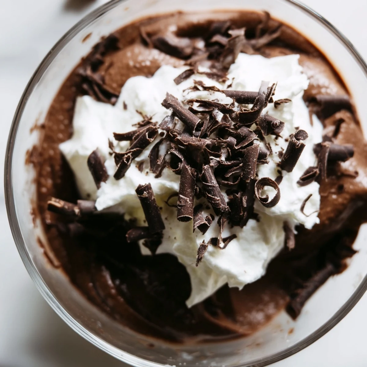 A close-up view of a single serving of chocolate mousse with whipped cream shavings, showcasing the airy texture and glossy chocolate surface against a rustic background.