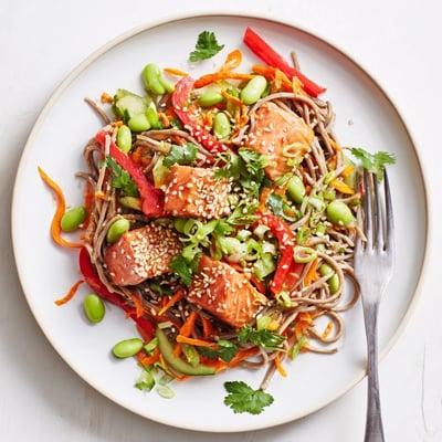 A close-up of Teriyaki Salmon Soba Noodle Salad shows flaky salmon, colorful bell peppers, and edamame tossed in tangy sesame dressing.